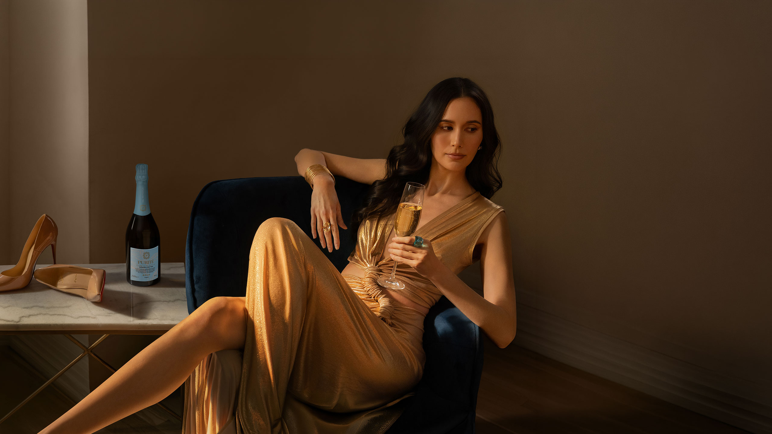 A woman in gold dress relaxing in a blue velvet chair. She is sipping a glass of Purité Sparkling Tea Brut Vineyard Style. On the table beside her is a bottle of Purité Sparkling Tea Brut Vineyard Style and her designer shoes.